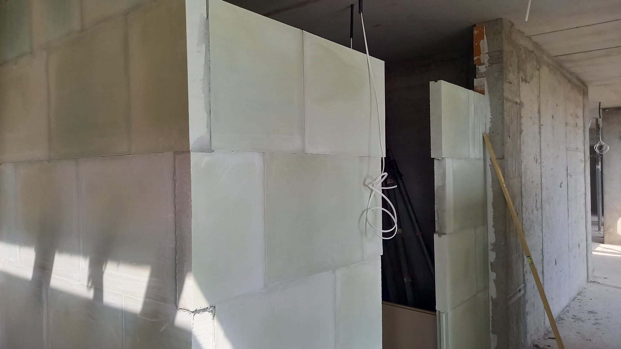 Wnętrze w trakcie prac wykończeniowych: ściany z płyt, widoczne łączenia, surowy beton w tle. Kabel zwisa z sufitu. Ujęcie z bliska, ukazujące detale konstrukcji.