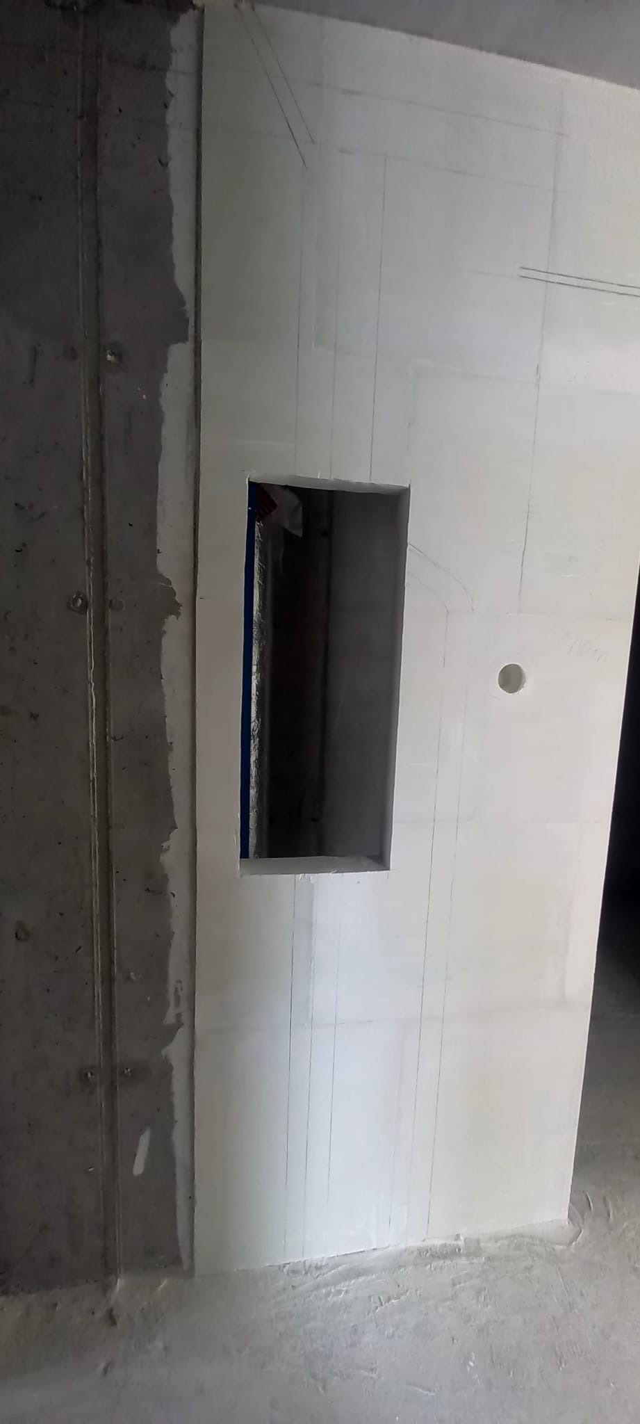 Ściana z otworem, przygotowana do dalszych prac wykończeniowych. Widoczne pionowe linie i surowy beton obok. Otwór zabezpieczony taśmą malarską.