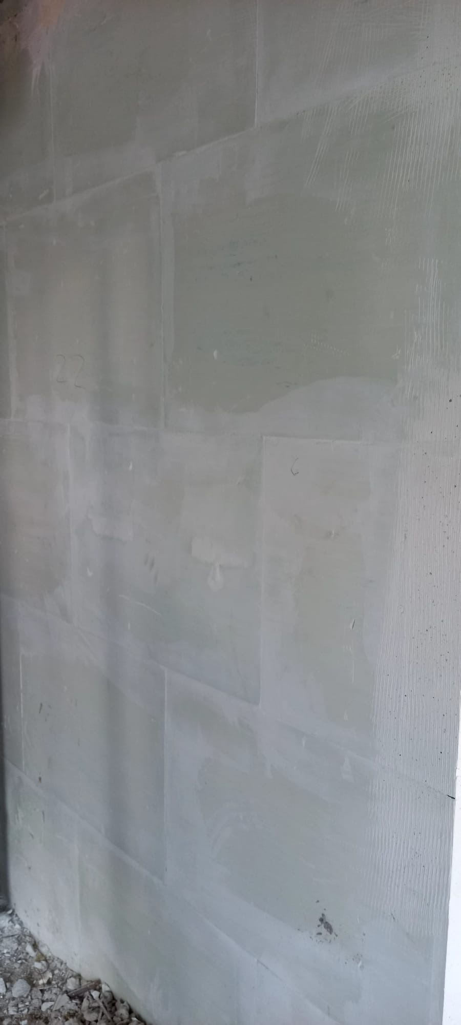 Ściana z szarej płyty kartonowo-gipsowej, częściowo wyszpachlowana, z widocznymi śladami po szpachlowaniu i numerem '22' napisanym na powierzchni. U dołu widoczne gruz.