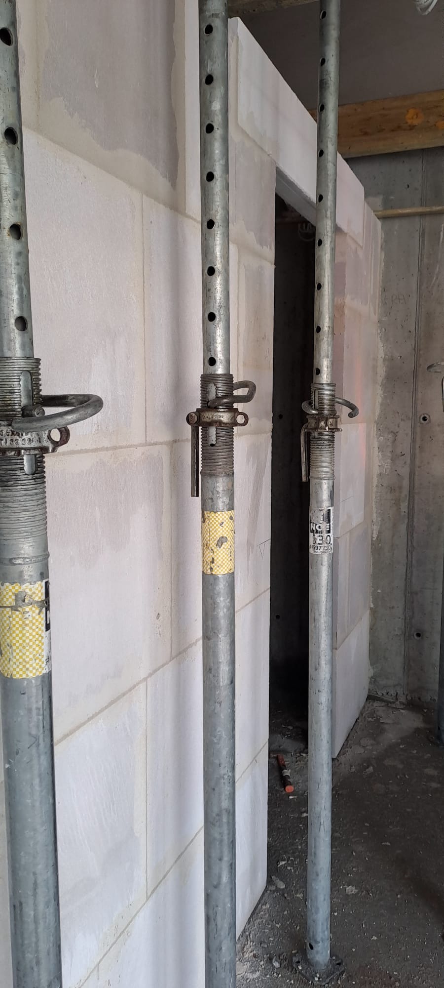 Wznoszenie ściany z bloczków z betonu komórkowego, podpartej metalowymi stemplami budowlanymi. Widoczne spoiny i niedoskonałości, surowy stan budowy.