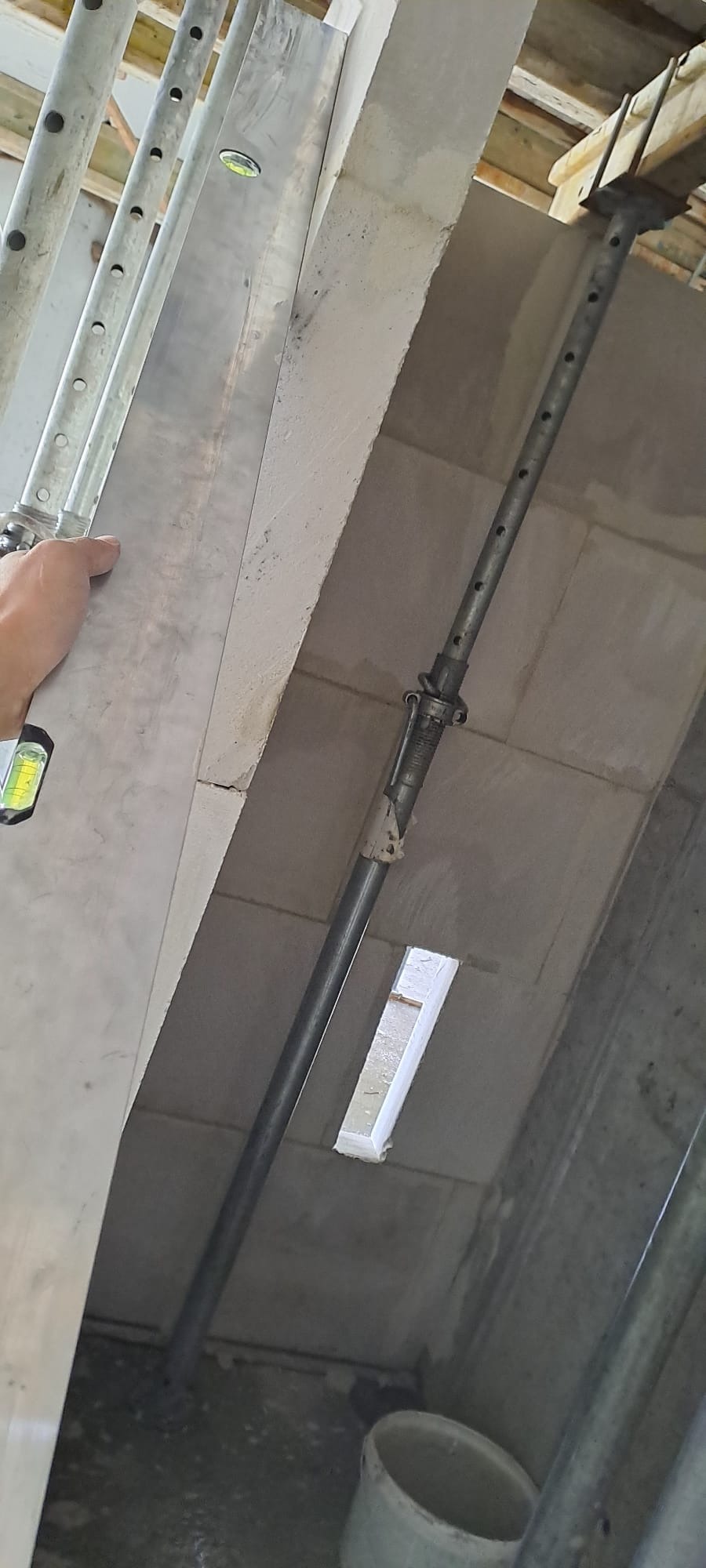 Wznoszenie ściany z betonu komórkowego, podpora stalowa, poziomica w użyciu. Widoczny fragment stropu i pustak wentylacyjny. Praca w toku, surowy stan.