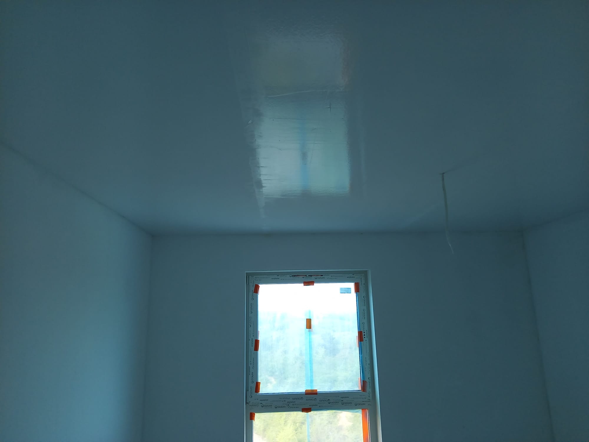 Wnętrze pokoju po świeżym malowaniu na biało, z oknem zabezpieczonym folią. Widoczne refleksy światła na gładkiej powierzchni sufitu i ścian.