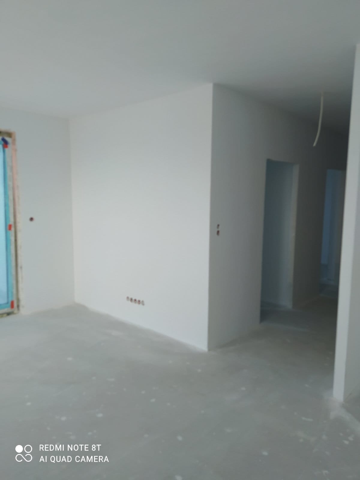 Wnętrze po gładziowaniu ścian, widoczne gniazdka elektryczne, surowa podłoga i przygotowane otwory drzwiowe. Ściany pomalowane na biało, w tle widoczne kolejne pomieszczenia.