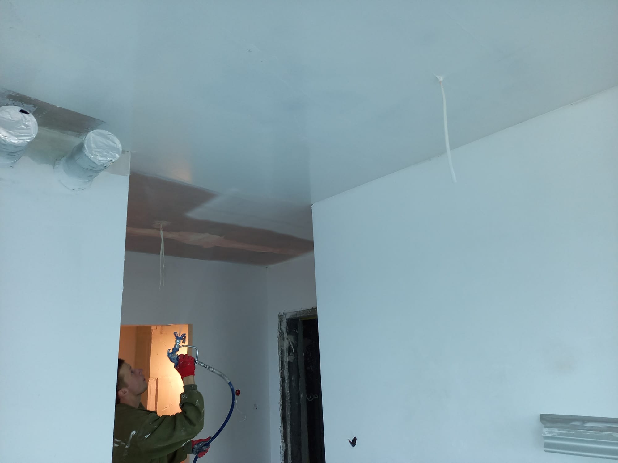 Pracownik w czerwonych rękawicach natryskowo nanosi białą farbę na sufit w pomieszczeniu. Widoczne zabezpieczone folią rury wentylacyjne i fragment ściany.