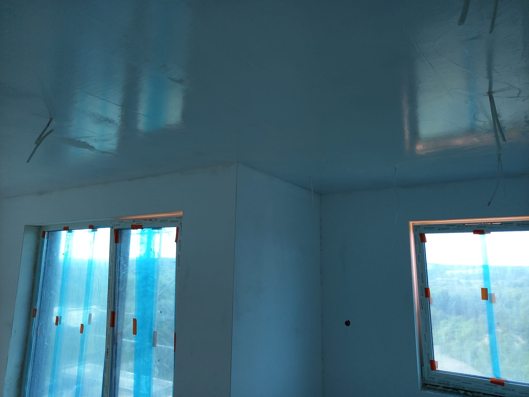 Pokój po gładziowaniu: gładka, błyszcząca powierzchnia sufitu odbija światło. Okna zabezpieczone folią. Widoczne ślady po taśmach malarskich. Ściany przygotowane do malowania.