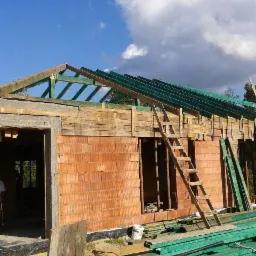 Wznoszenie ścian i konstrukcji dachu domu z czerwonej cegły, widoczne drewniane elementy konstrukcyjne, drabina oparta o ścianę, błękitne niebo w tle.