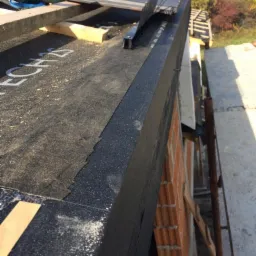 Widok z góry na fragment dachu w trakcie prac: czarna membrana z widocznym napisem, narzędzia, elementy konstrukcyjne, ceglany komin i rusztowanie. W tle zieleń drzew i zabudowania.