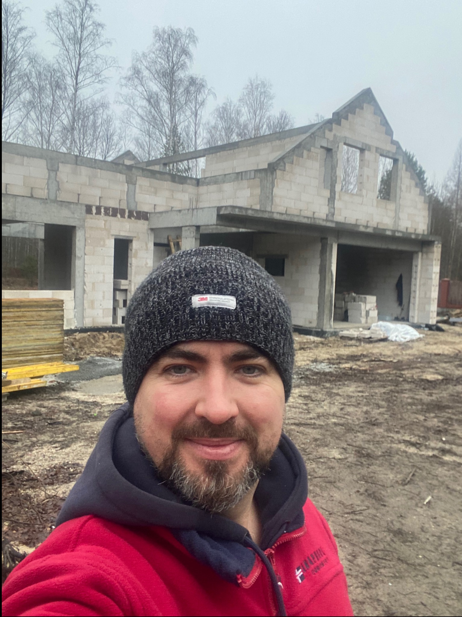 Mężczyzna w czapce na tle surowego stanu domu z betonu komórkowego, widoczne drewniane stemple i pustaki. Budowa w toku, pochmurny dzień.