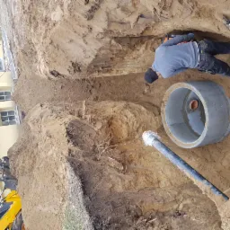 Pracownik w wykopie w Szczecinie układa betonowy krąg kanalizacyjny; widoczna koparka i budynek w tle.