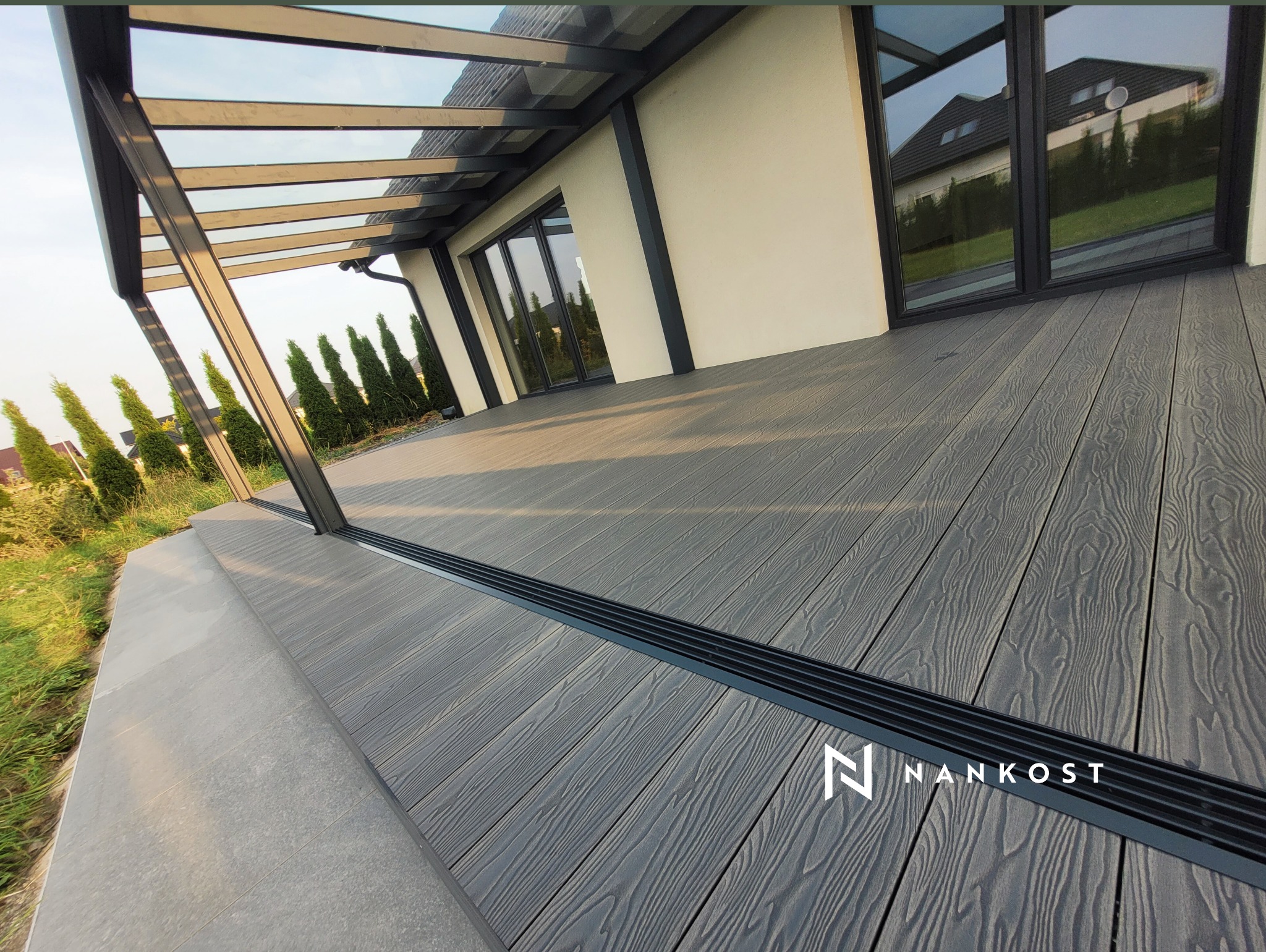 Szare deski tarasowe kompozytowe z widocznym systemem odprowadzania wody i zadaszeniem, na tle domu z dużymi oknami.