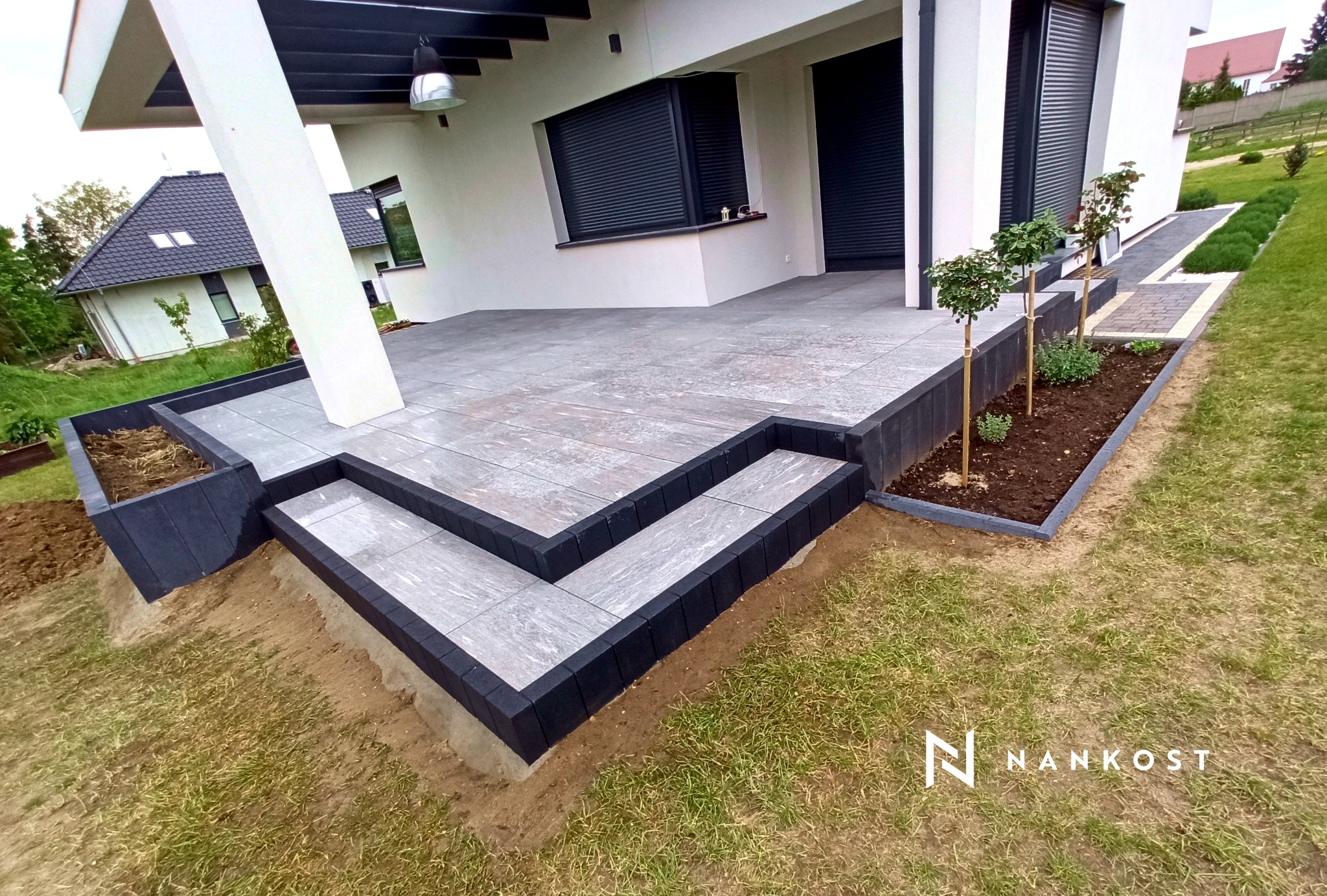 Nowoczesny taras z szarych płyt betonowych, obrzeża z czarnej kostki, schody tarasowe, minimalistyczna architektura domu z białymi ścianami i szarymi roletami, rabaty z młodymi drzewkami, trawnik.