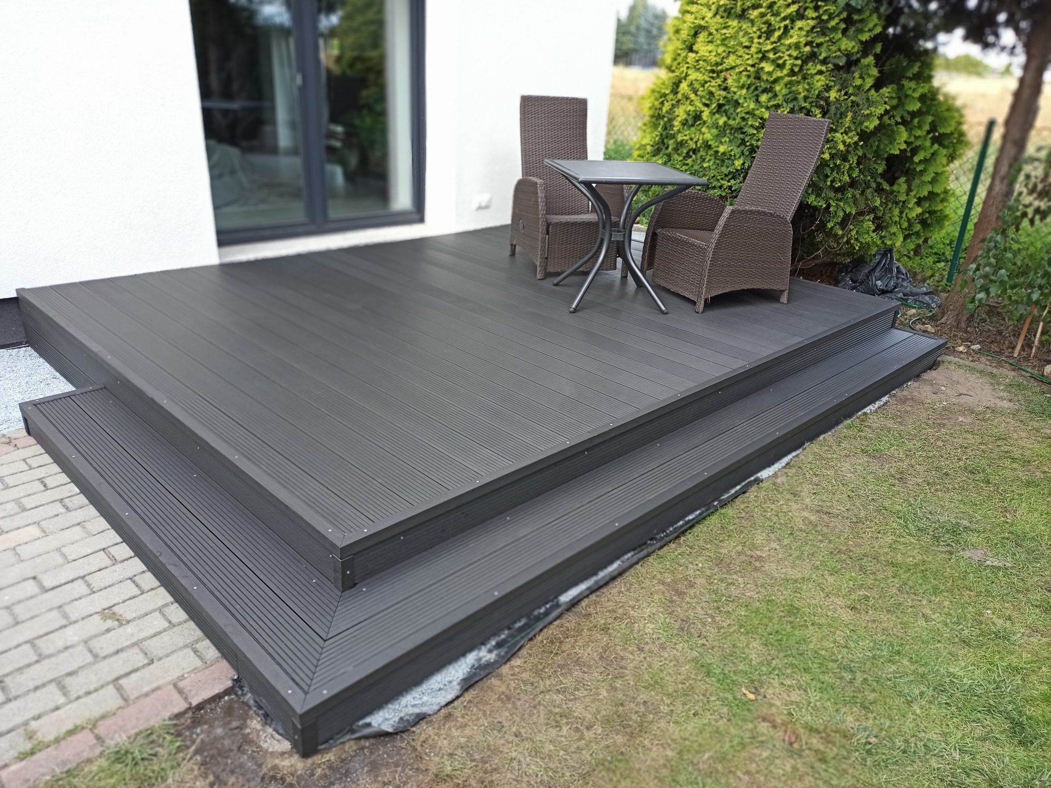 Nowoczesny taras kompozytowy w kolorze grafitowym z dwoma stopniami, wyposażony w zestaw mebli ogrodowych z plecionki technorattanowej, na tle białej ściany budynku i zieleni ogrodu.