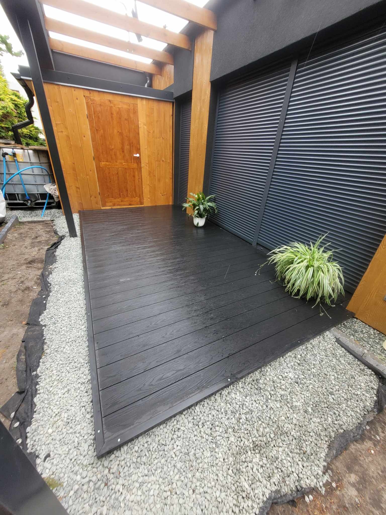 Nowoczesny taras kompozytowy w Jełowej z czarnych desek, otoczony białym żwirem i roślinami w donicach, przylegający do drewnianej konstrukcji i ciemnej ściany z żaluzjami.