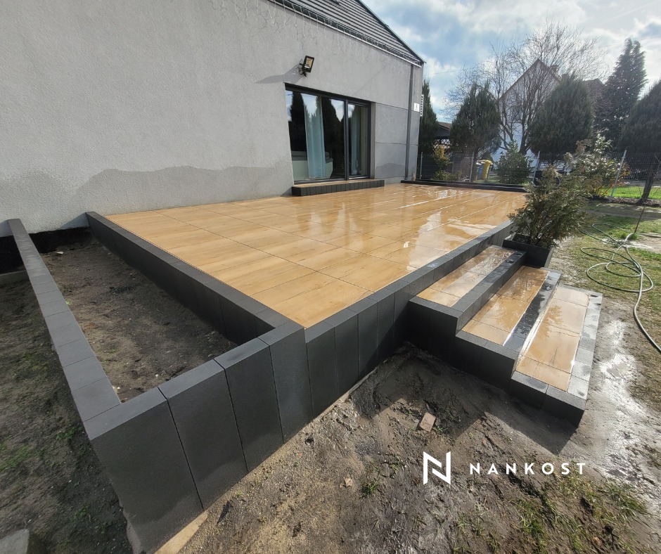 Nowoczesny taras z płytek betonowych imitujących drewno, obrzeża z grafitowych bloczków, trzy stopnie wejściowe, widok na dom i ogród w pochmurny dzień.