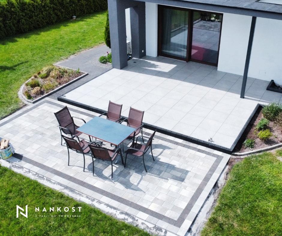 Nowoczesny taras z jasnoszarych płyt betonowych, z ciemnoszarą obwódką, wyposażony w stół i krzesła z metalową ramą, widok z góry na tle domu z dużymi przesuwnymi drzwiami i zielonego trawnika.