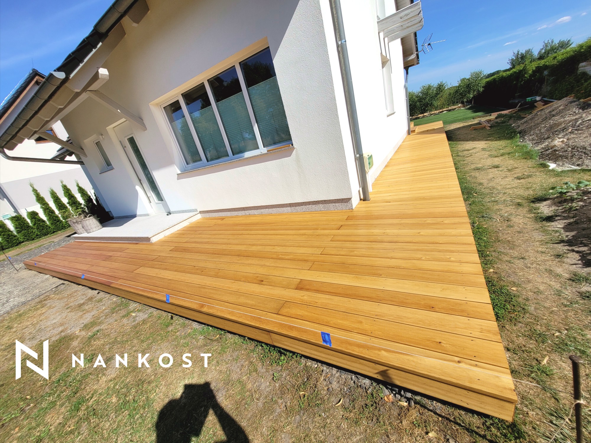 Świeżo wykończony drewniany taras przylegający do białego domu z widocznymi niebieskimi taśmami zabezpieczającymi na krawędziach, z logo firmy Nankost w lewym dolnym rogu.