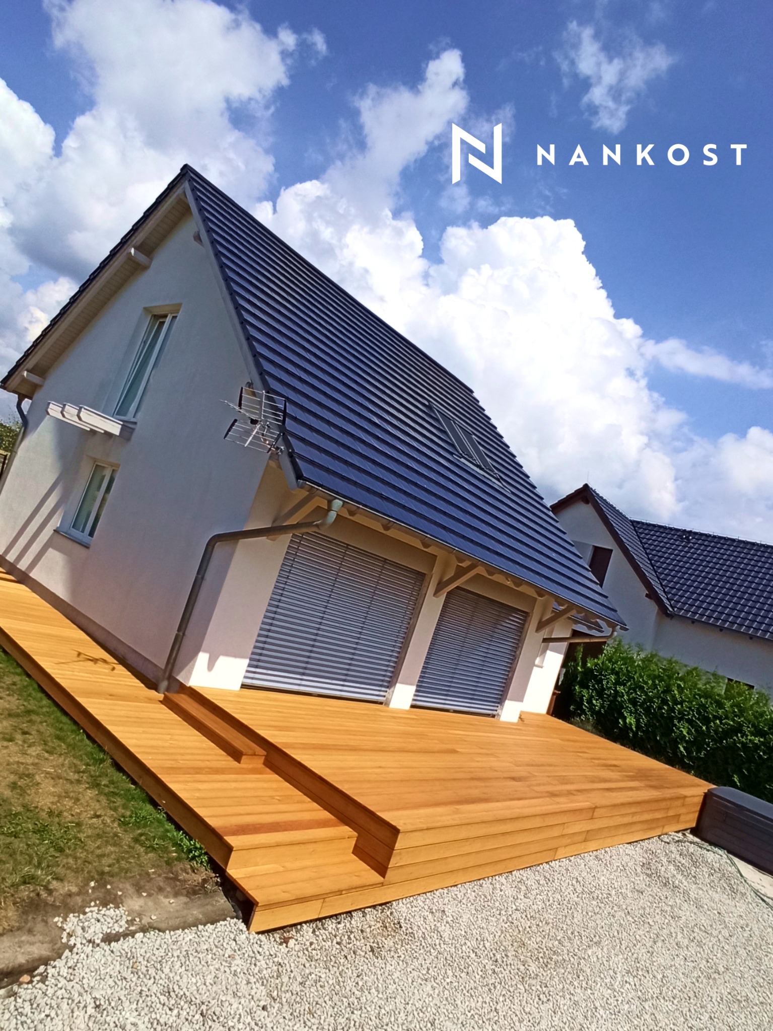 Nowoczesny dom z jasną elewacją i drewnianym tarasem na dwóch poziomach, z widocznym logo firmy NANKOST na tle błękitnego nieba z obłokami.