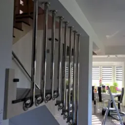 Nowoczesna balustrada schodowa ze stali nierdzewnej, z pionowymi słupkami mocowanymi do stopni, widok na jasne wnętrze domu z jadalnią w tle.