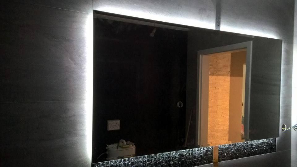 Prostokątne lustro z podświetleniem LED umieszczone na szarej ścianie wyłożonej płytkami, z dekoracyjnym pasem mozaiki u dołu i widocznym fragmentem instalacji elektrycznej.