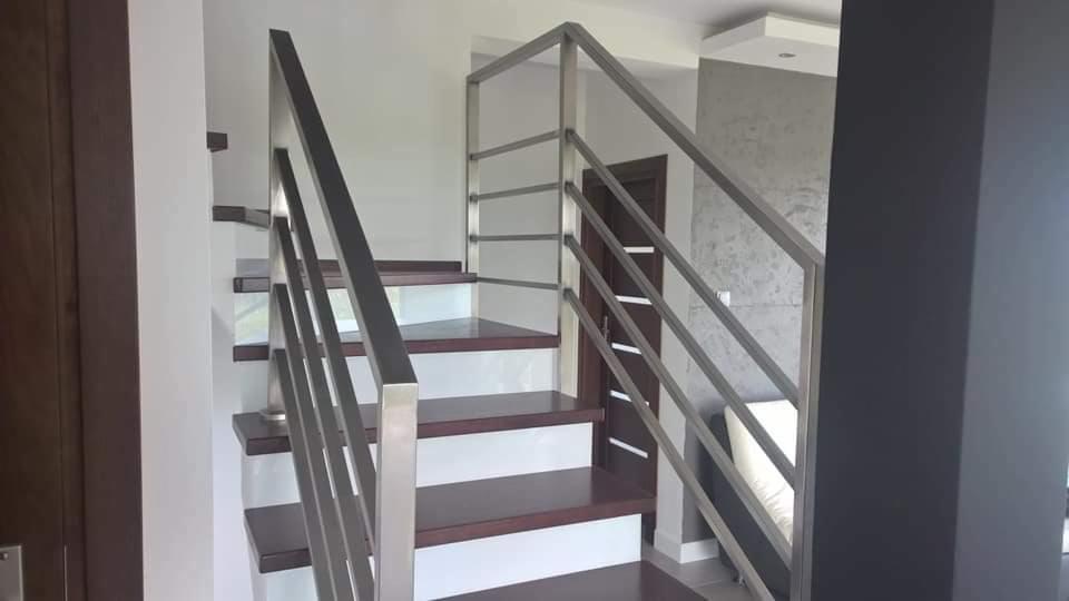 Nowoczesne schody z drewnianymi stopniami i minimalistyczną, metalową balustradą ze stali nierdzewnej, widok z dołu na tle białych ścian i brązowych drzwi.
