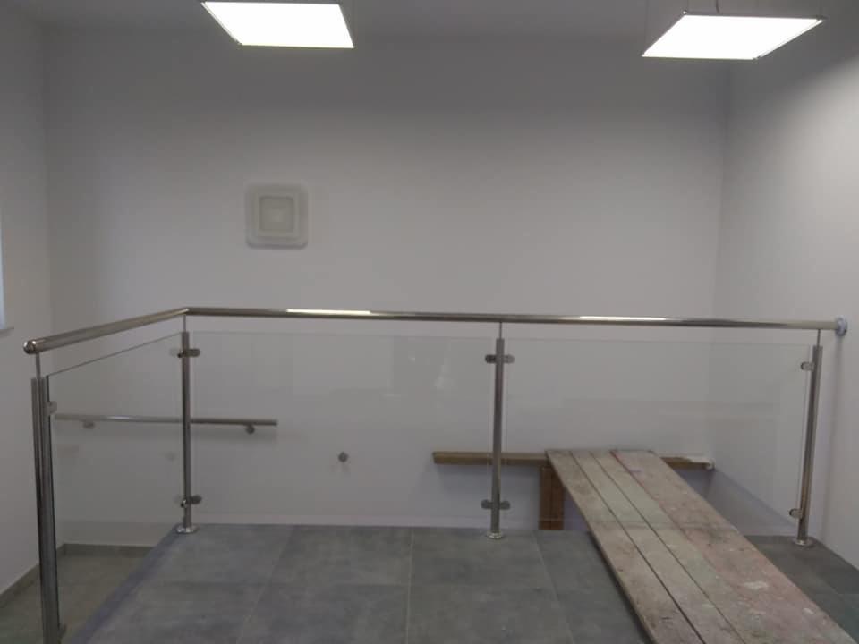 Szklana balustrada z metalowymi słupkami i poręczą ze stali nierdzewnej, widoczna deska podestowa na szarych płytkach podłogowych, minimalistyczne wnętrze z białymi ścianami i kwadratowymi lampami...