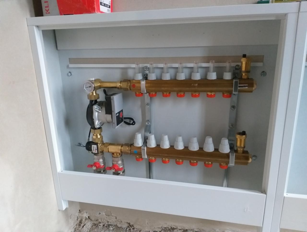 Rozdzielacz ogrzewania podłogowego w białej szafce, z pompą obiegową i zaworami, widok z bliska na instalację w trakcie montażu.