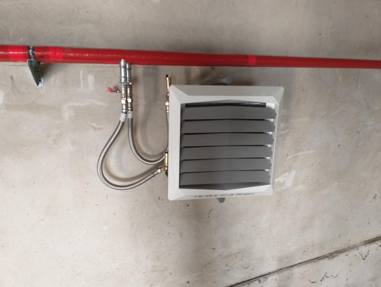 Nagrzewnica wodna Volcano zawieszona na ścianie, podłączona do rury grzewczej za pomocą elastycznych przewodów w oplocie stalowym, surowy beton w tle.