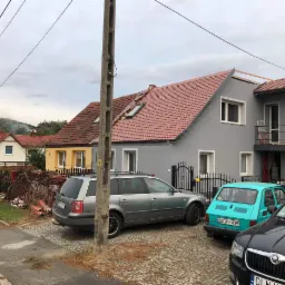 Szary dom jednorodzinny z czerwoną dachówką w trakcie remontu, widoczne rusztowania na dachu, na podwórku zaparkowane samochody, w tym turkusowy Fiat 126p.