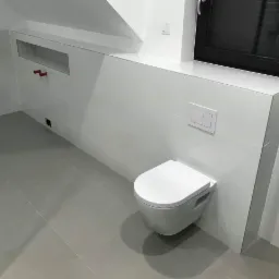 Nowoczesna, minimalistyczna toaleta podwieszana z białym panelem spłukującym, zamontowana na ścianie z białych płytek w łazience w Kamiennej Górze. Widoczne szare płytki podłogowe i wnęka w ścianie.