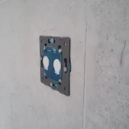 Szary panel sterujący inteligentnego domu, z widoczną elektroniką, zamontowany na szarej, teksturowanej ścianie.