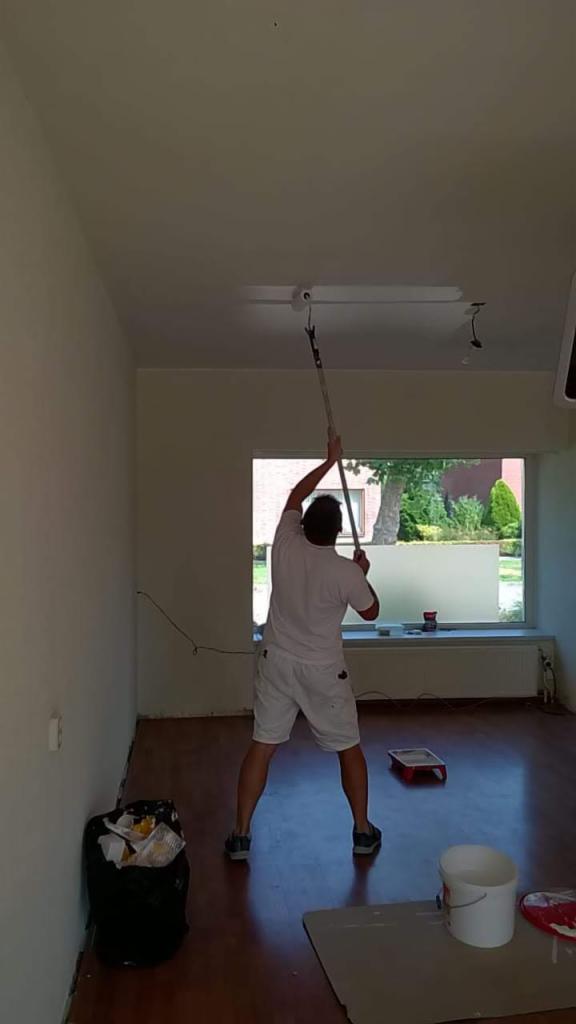 Mężczyzna maluje sufit pomieszczenia wałkiem na długim kiju, widoczny kubeł z farbą, kuweta malarska i śmieci w czarnym worku.