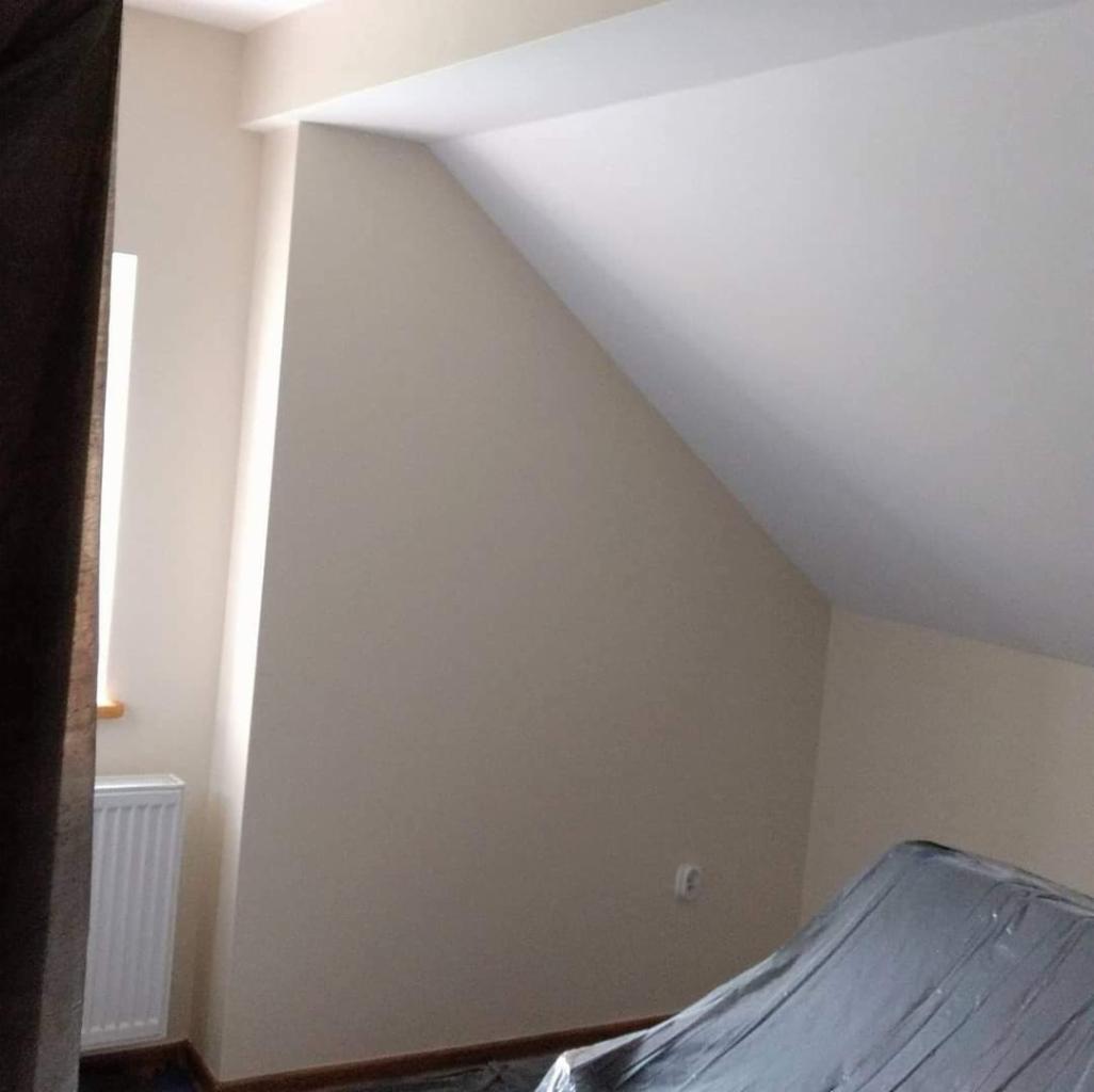 Pomalowane na beżowo poddasze ze skosami, zabezpieczone meble folią, widoczny grzejnik i fragment okna.