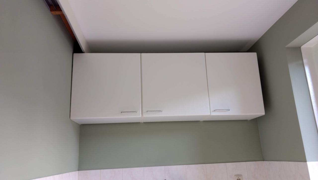 Białe szafki kuchenne wiszące pod sufitem, zamontowane w pomieszczeniu z zielonymi ścianami i białą glazurą