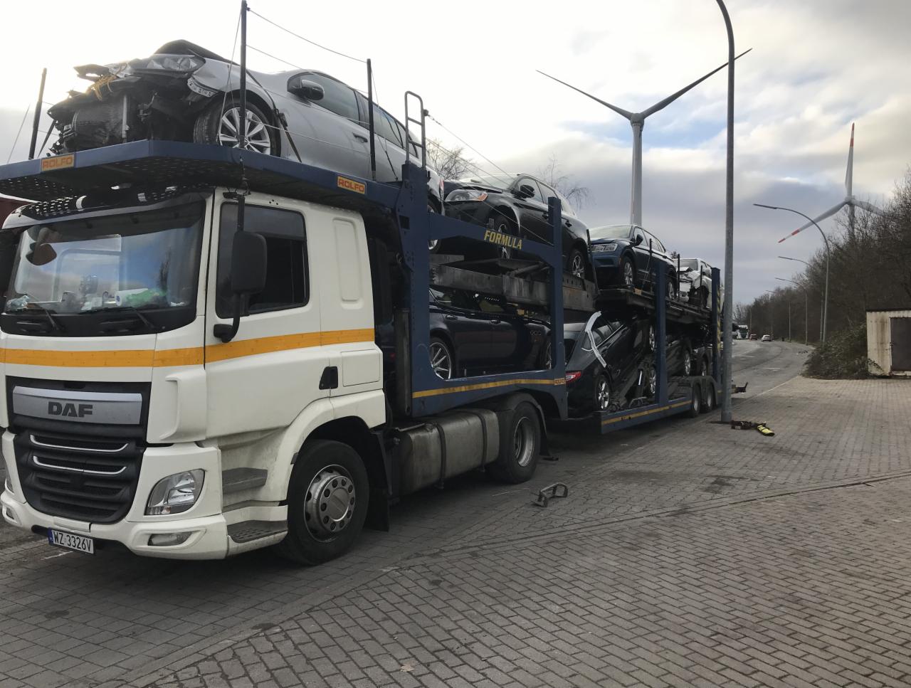 Laweta DAF z uszkodzonym srebrnym samochodem na górnym pokładzie oraz kilkoma innymi autami na pozostałych poziomach, zaparkowana na brukowanym placu z turbinami wiatrowymi w tle.