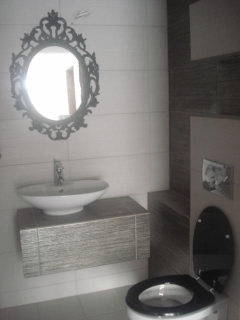 Nowoczesna łazienka z owalną umywalką nablatową, szafką pod umywalką wykończoną płytkami imitującymi drewno, ozdobnym lustrem w czarnej ramie i toaletą z czarną deską.