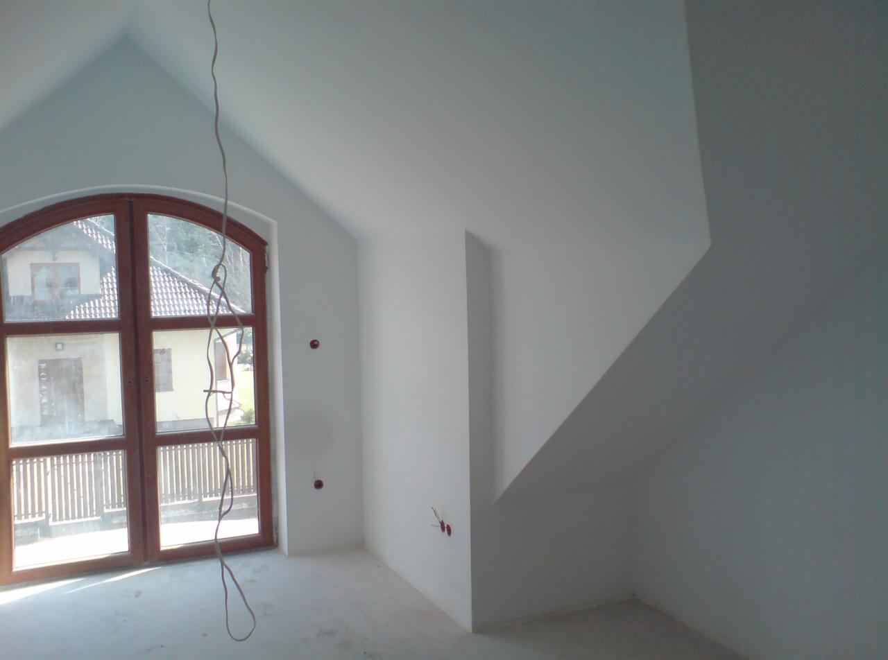 Poddasze w trakcie prac wykończeniowych: białe ściany, drewniane okno z widokiem na dom i ogród, wystające kable elektryczne ze ściany.