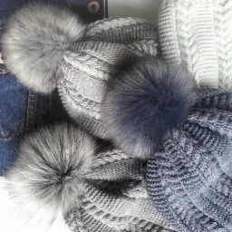 Stos dzianinowych czapek z futrzanymi pomponami w odcieniach szarości i błękitu, ułożonych obok dżinsowej kurtki z metalowym guzikiem.