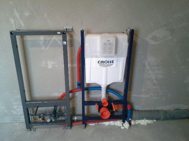 Stelaż podtynkowy WC marki Grohe z podłączonymi rurami wodnymi (czerwone i niebieskie) i kanalizacyjnymi, zamontowany przy surowej, nieotynkowanej ścianie.
