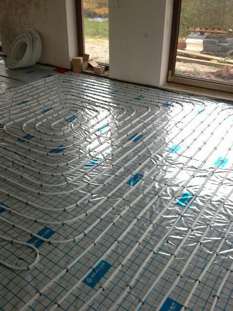Białe rury układane w pętle na podłodze z siatką i folią, przygotowanie do wylewki pod ogrzewanie, widoczne okno i zwój rur.
