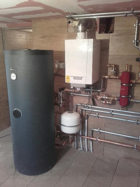 Bojler i piec gazowy z podłączeniami hydraulicznymi i rurami w surowym pomieszczeniu z płytami OSB na ścianach i suficie.