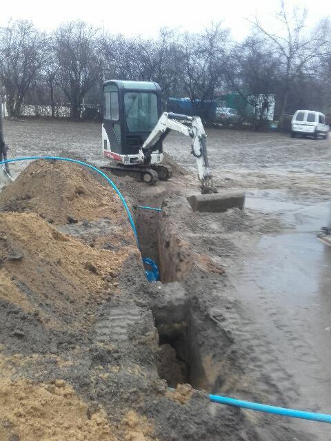 Mini koparka Bobcat przy wykopie z widoczną niebieską rurą w ziemi, w tle drzewa bez liści i biały bus.