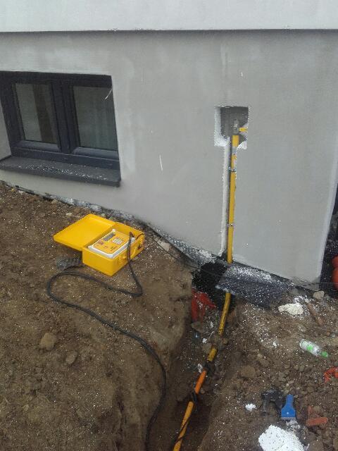 Żółta rura gazowa w wykopie przy fundamencie budynku, podłączona do ściany z otworem. Żółty miernik w otwartej walizce obok wykopu.