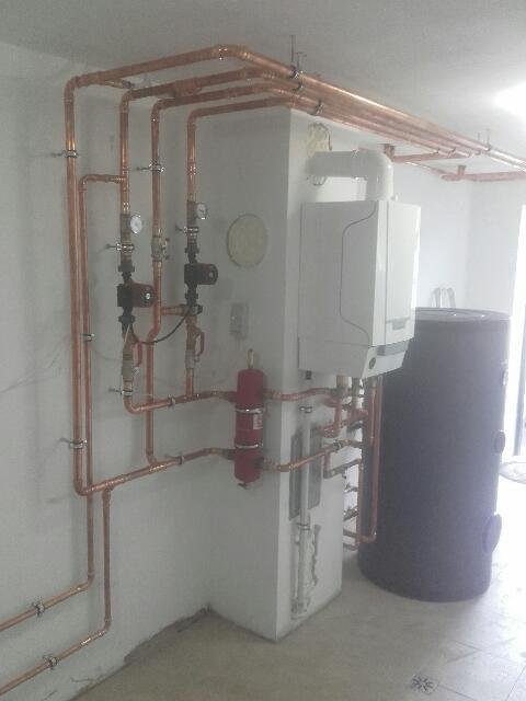 Nowoczesny piec grzewczy z podłączonym zasobnikiem ciepłej wody i rozbudowaną instalacją miedzianą, zamontowany w pomieszczeniu gospodarczym.