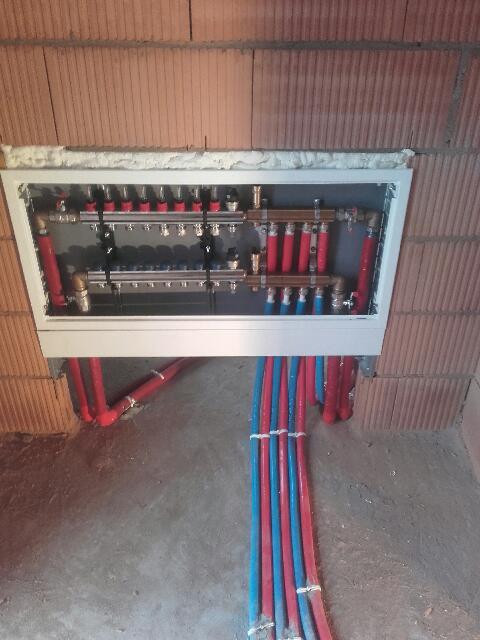 Rozdzielacz ogrzewania podłogowego w zabudowie, widoczne czerwone i niebieskie rury prowadzące do rozdzielacza, tło z cegły.