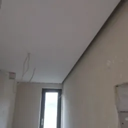 Wnętrze w trakcie prac wykończeniowych: widok na sufit po nałożeniu gładzi, okno z widocznym światłem dziennym oraz stos płyt kartonowo-gipsowych opartych o ścianę.