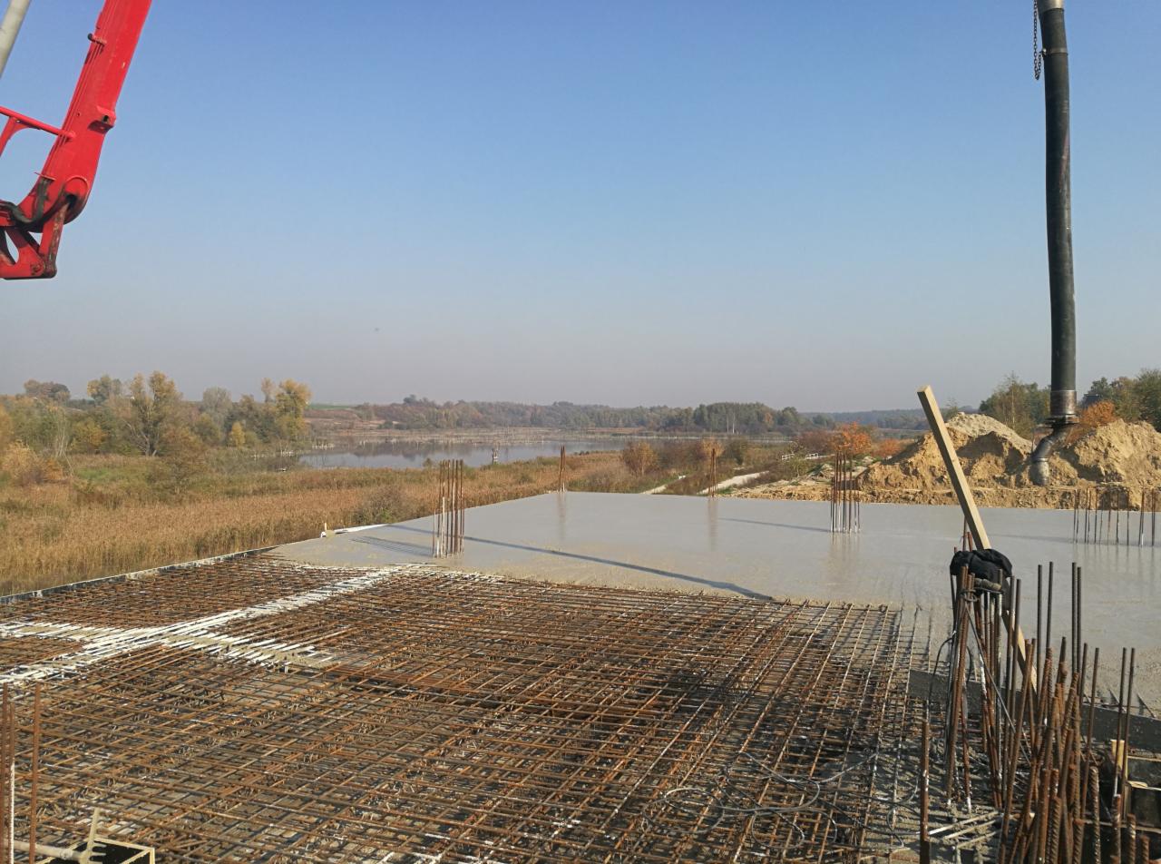 Wylewanie betonu na zbrojone fundamenty budynku z widokiem na jezioro, w tle las i błękitne niebo.