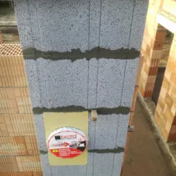 Wznoszona ściana z szarego bloczka, z widocznymi spoinami zaprawy, z fragmentem reklamy kominów 'Brata' przymocowanej do ściany, w tle fragmenty konstrukcji domu z czerwonej cegły dziurawki.