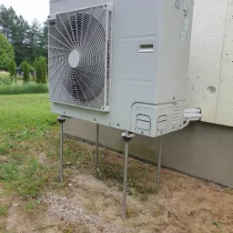 Zewnętrzna jednostka klimatyzacji typu monoblok, zamontowana na metalowych nóżkach przy ścianie budynku ocieplonego styropianem. Widoczne rury wychodzące ze ściany i podłączone do urządzenia.