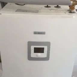 Biały, nowoczesny piec gazowy marki Bosch z szarym panelem sterowania, widoczne rury i zawory z izolacją termiczną w tle, u dołu koszyk z rzeczami.