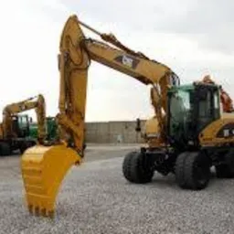 Żółta koparka kołowa Caterpillar z łyżką opuszczoną do ziemi na żwirowym placu, w tle druga koparka i betonowy mur.